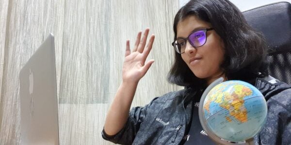 A girl sitting at a desk with a laptop open, waving to the camera. On the desk, there is a small globe.