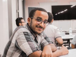 Abdulrahman sitting at a table with his arms on the desk, and two people in the background