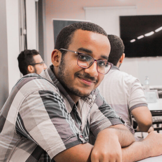 Abdulrahman sitting at a table with his arms on the desk, and two people in the background