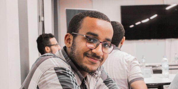 Abdulrahman sitting at a table with his arms on the desk, and two people in the background