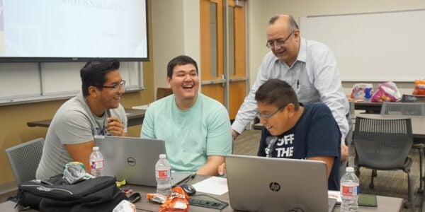 Three students working on laptops at a desk with a teacher behind them