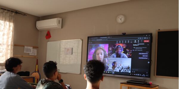 Three students sitting at desks in a classroom, facing a large screen displaying a Zoom meeting with other students.